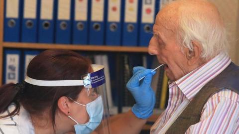 Healthcare worker wearing a visor and face mask taking a mouth swab from an elderly man during the Covid-19 pandemic