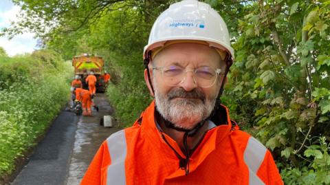 A man in an orange safety jacket with white hard hat. He has glasses and a grey beard. Behind him is a team of pothole repair workers with a lorry