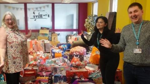 Two women and one man stood next to a large pile of Christmas presents. All three people have their hands in the air and are smiling, and looking directly at the camera. 