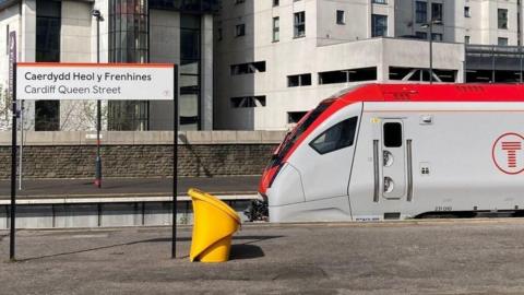 A Transport for Wales train pulled into Cardiff Queen Street, with the red T logo in a circle displayed on the side of the grey and red train. There is a gritting bin in shot, and a sign for Cardiff Queen Street, and its Welsh version Caerdydd Heol y Frenhines, at the left hand side.