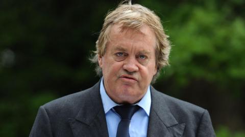The Duke of Marlborough faces the camera, not smiling, in an outdoor location. He wears a dark grey suit, light blue shirt and dark blue tie, and has fair, slightly untidy, hair.