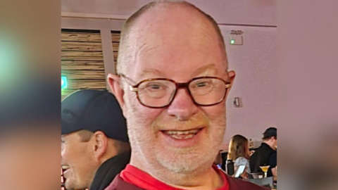 A man wearing glasses and a red shirt smiles at the camera, with other people seated at tables in a softly lit indoor setting behind him.