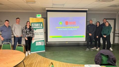 Seven people are pictured next to a screen which says "Leicestershire and Rutland County FA Mental Health Friendly Clubs"