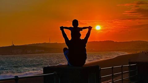 A silhouetted man holds his little boy in the air as the sun sets in the distance. 