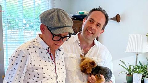 Dr. Trevor Yellon and his late partner Drew. They are both wearing white shirts and Dr. Trevor is holding a dog. 