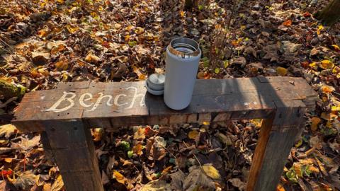 An opened flask sits on a small wooden bench which has the word "bench" written on it. Brown leaves can be seen covering the ground underneath with shadows of trees.