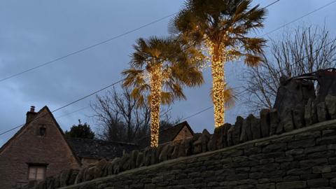 Two large palm trees, the trunks of which are entirely covered with fairy lights, stick out above a traditional stone wall. The pointed roof of the house can be seen jutting above the wall. 