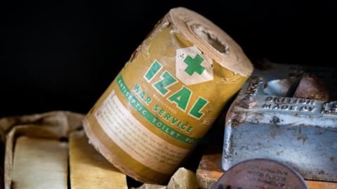 An old-looking roll of Izal toilet paper, the type that would have been used by the military in WW1. The paper cover around the roll says "Izal, Registered, War Service, Antiseptic Toilet Roll".