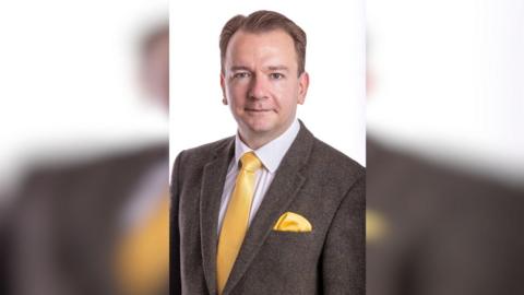 A man in a suit. He is wearing a yellow tie, a white shirt and a brown suit. The background is white. 