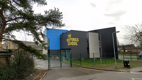 The school is a modern box-like building clad in black and bright blue, with The Deepings School split across one side of the building on a 45 degree angle in yellow capital letters.  The photo is taken on an overcast day.