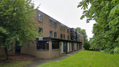 A three‑storey brick residential building with rows of windows and small balconies, set beside a paved walkway and surrounded by grass and leafy trees on a quiet, green campus‑like setting.