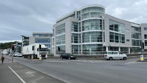 A grey building with blue windows with a traffic light controlled junction in front. An articulated lorry is waiting at the lights and two cars are turning left into Admiral Park. 