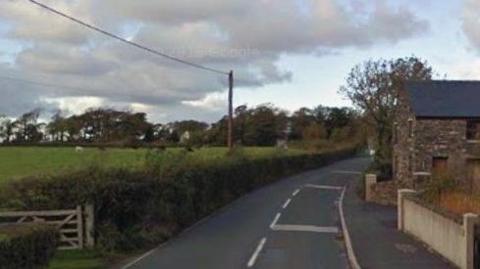 The road leading away from the junction with St Judes. There is a green field and hedge on the left and a wall and stone built building on the right.