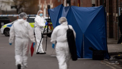 A forensic crime scene showing officers in white PPE and walking towards a blue police tent.