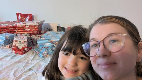 A pile of gifts on a table behind Harper, 8, on the left and Kirsty on the right. Kirsty is taking a selfie and they are both smiling at the camera. The presents are piled up and are in red, blue and white Christmas presents.