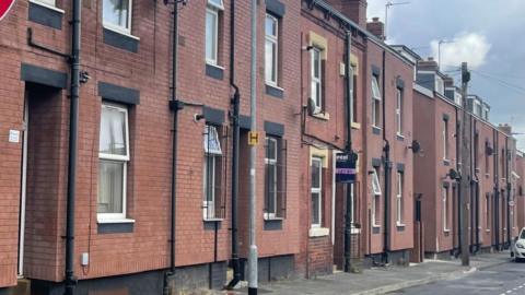 Terraced redbrick houses, Some with black grills over the windows. One has a purple 'Let By' sign at the front.