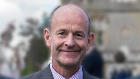 A man wearing a jacket and striped shirt is smiling at the camera. The background is blurred.