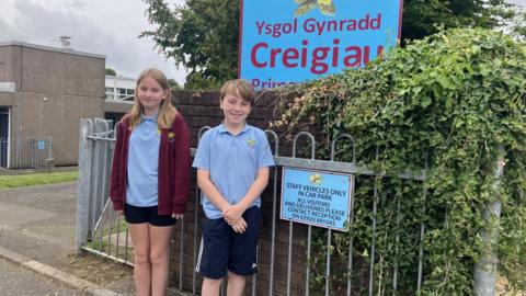 Rua and Ella in front of the school gates of Ysgol Gynradd Creigiau. They are wearing light blue uniform polo shirts with the school's logo on and shorts. 