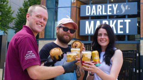 Two men and a woman hold up beers and pies outside Darley Street Market.