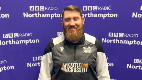 A man with short brown hair brushed to the left with a big beard stands in front of the purple BBC Radio Northampton branded wall smiling at the camera. He is wearing a black, white and grey zipped top with "The Man Cave" written above the left side of his chest.