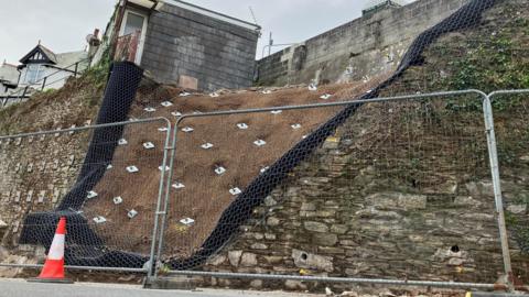 Temporary security fencing in front of a stone wall where the middle section has been replaced with heavy metal ground retaining pins at about 8m deep covered by heavy wire.  