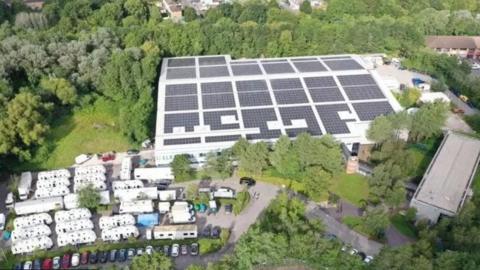 An aerial view of Bottle Yard studios showing a large warehouse roof covered in solar panels and surrounded by trees, there appears to be seven rows of trailers in the bottom left corner of the frame.