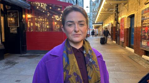 Ros Morgan, wearing a purple jacket and floral scarf, looks at the camera. SHe has short hair and a neutral expression
