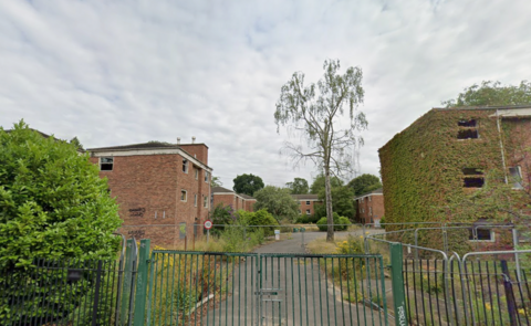 Derelict three-storey halls of residence buildings, one of which is covered in behind fencing ivy. The buildings are behind silver fencing and a locked green gate.