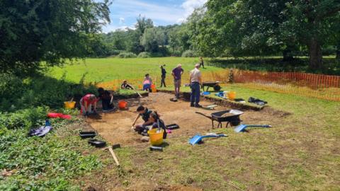 A team of archaeologists works in a hole in a grassy field. It is sunny. 