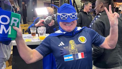 Man in a blue facemask and cape round his neck. He is wearing a Scotland strip which has the emblem for the Qatar world cup final on it next to the Argentina flag and French flag.