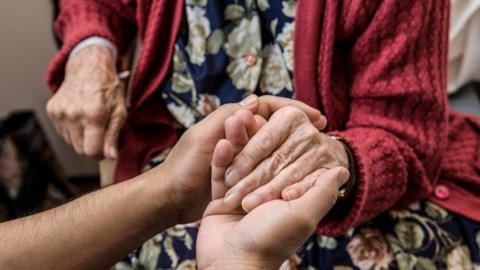 An elderly person in a red cardigan and floral dress has their hands gently held by another individual.