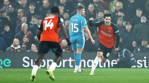 Liam Walsh (right) holds his fist up in celebration after scoring the winning goal. In the far left his teammate celebrates with him, they are both wearing the red Luton town home kit. Central a Northampton Town player (Evans) looks disappointed in light blue Northampton away kit. In the background, Northampton Town fans look equally disappointed. 