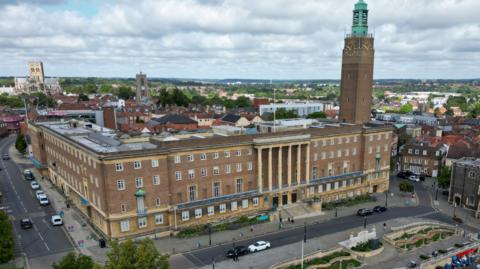 A drone image looks down on a four-storey city hall made of grey stone, featuring white windows and a balcony with columns, with a clock tower to the right of the building. It is shown surrounded by the trees and buildings of a city's urban sprawl, including a large cathedral on the horizon.