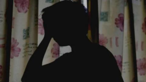 A silhouette of a person holding their head in one hand in a darkened room with the curtains drawn.