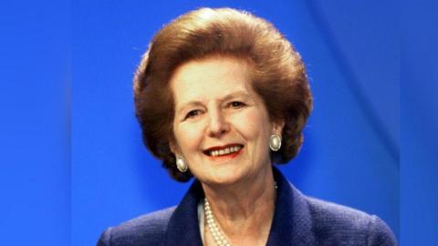 Margaret Thatcher has brown eyes and short brown voluminous hair swept backwards. She is wearing a cobalt blue collar-less blazer with a pearl necklace and a pearl-stud earrings and red lipstick. She is looking to the right and smiling with a blue background.