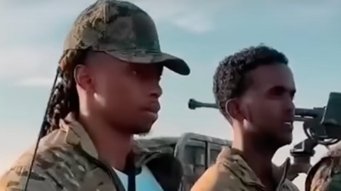 The Liverpool rappers wear combat clothing and are seen with assault rifles.