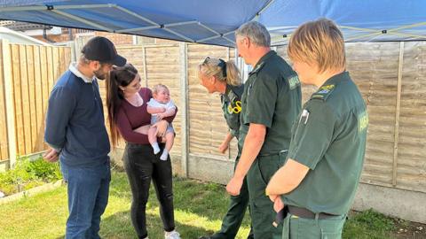 Three ambulance crew dressed in green uniform look at baby Oscar who is being held by his mother in the garden of the family home with his father Connor dressed in blue cap, shirt and jeans looking on