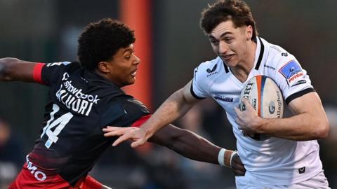Noah Caluori, wearing a black Saracens shirt, tackles Campbell Ridl in a white Exeter shirt.