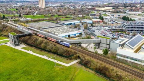 An aerial view of a modern railway station with a train moving along elevated tracks. The station has covered platforms and walkways on either side, with a pedestrian tunnel running beneath the tracks. Surrounding the station are industrial buildings, car parks, and nearby residential areas.