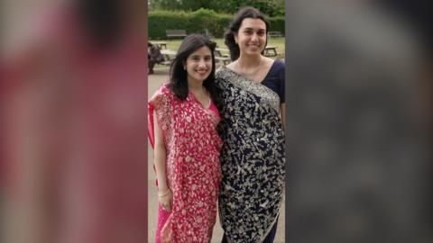 Two young women, both with long dark hair, stand side by side smiling. They are both wearing intricately-patterned saris.