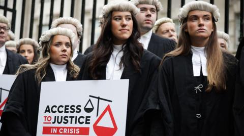 Three female barristers in white wigs and black gowns, holding a sign which reads Access to Justice in Crisis.