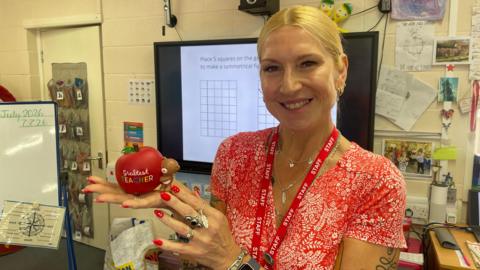 Teacher Shelley Grosse holding an ornament with the wording "Greatest Teacher". She is wearing a red summer dress and has tied back blonde hair. She has a number of tattoos on her left arm and a watch on her wrist.