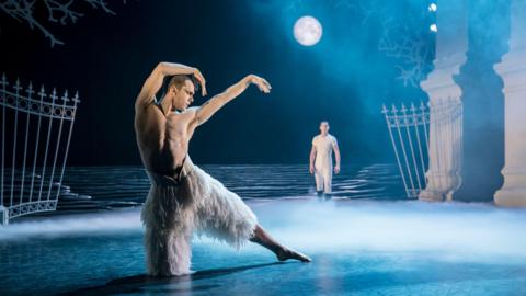 A scene from Swan Lake. A male dancer rests on one knee with his left leg outstretched and his arms curled around his head. He is bare chested and wears white feather trousers. Behind him is a man in a white T-shirt and trousers standing looking at him under a full moon. The set features wrought iron gates and part of a large stone building partly obscured by a low mist.