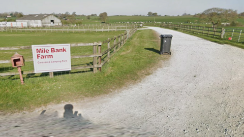 The entrance to a caravan park on farmland with a red-on-white sign reading Mile Bank Farm Caravan and Camping Park, next to a hardcore driveway edged by a wheelie bin and wooden fences, with fields of cattle in the distance.