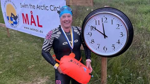 Roz Rapp standing in a wet suit holding a yellow float next to a clock with the time 11:50pm on the clock. She is smiling with medals around her neck and is in front of a sign which says "swim the arctic circle".