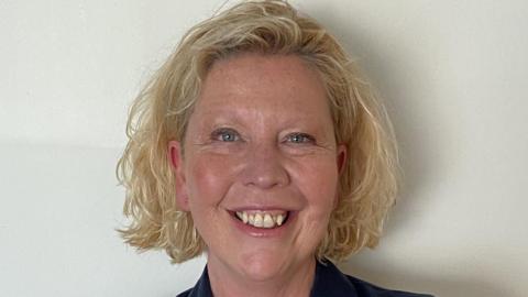 A photo of a lady with blonde hair. She is wearing a navy jacket and smiling at the camera.