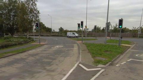 A large road junction with carriageways turning left and right, split by a triangular grass verge and crossing points. Four sets of traffic lights can be seen, all on green. A tree stands to the left. A white van is waiting at lights in the background. The sky is grey.