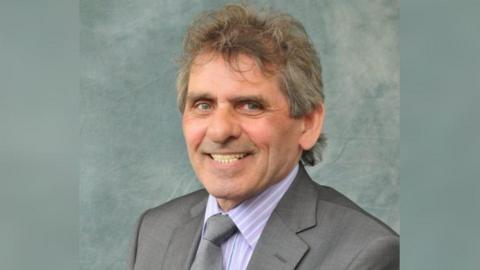 Denny Wilson is smiling at the camera. He is wearing a grey suit jacket , a grey tie and a shirt with yellow and lilac stripes. He had blue eyes and grey hair.