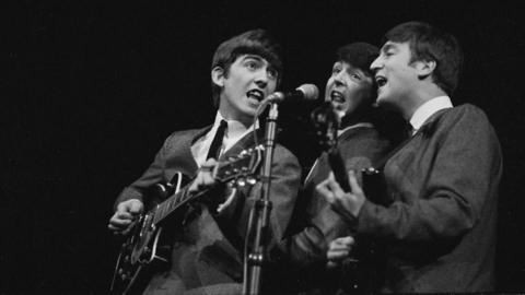A black and white image showing three members of The Beatles - from left to right George Harrison, Paul McCartney and John Lennon - sharing a microphone as they perform on stage against a black background. They are wearing suits, white shirts and black ties.