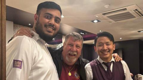 Professor Carl Chinn with employees Hamza Hussain and Bishal Tamang. They are standing either side of him wearing their purple work uniforms. Everyone is smiling. Carl Chinn has an Aston Villa top on and is wearing a necklace.